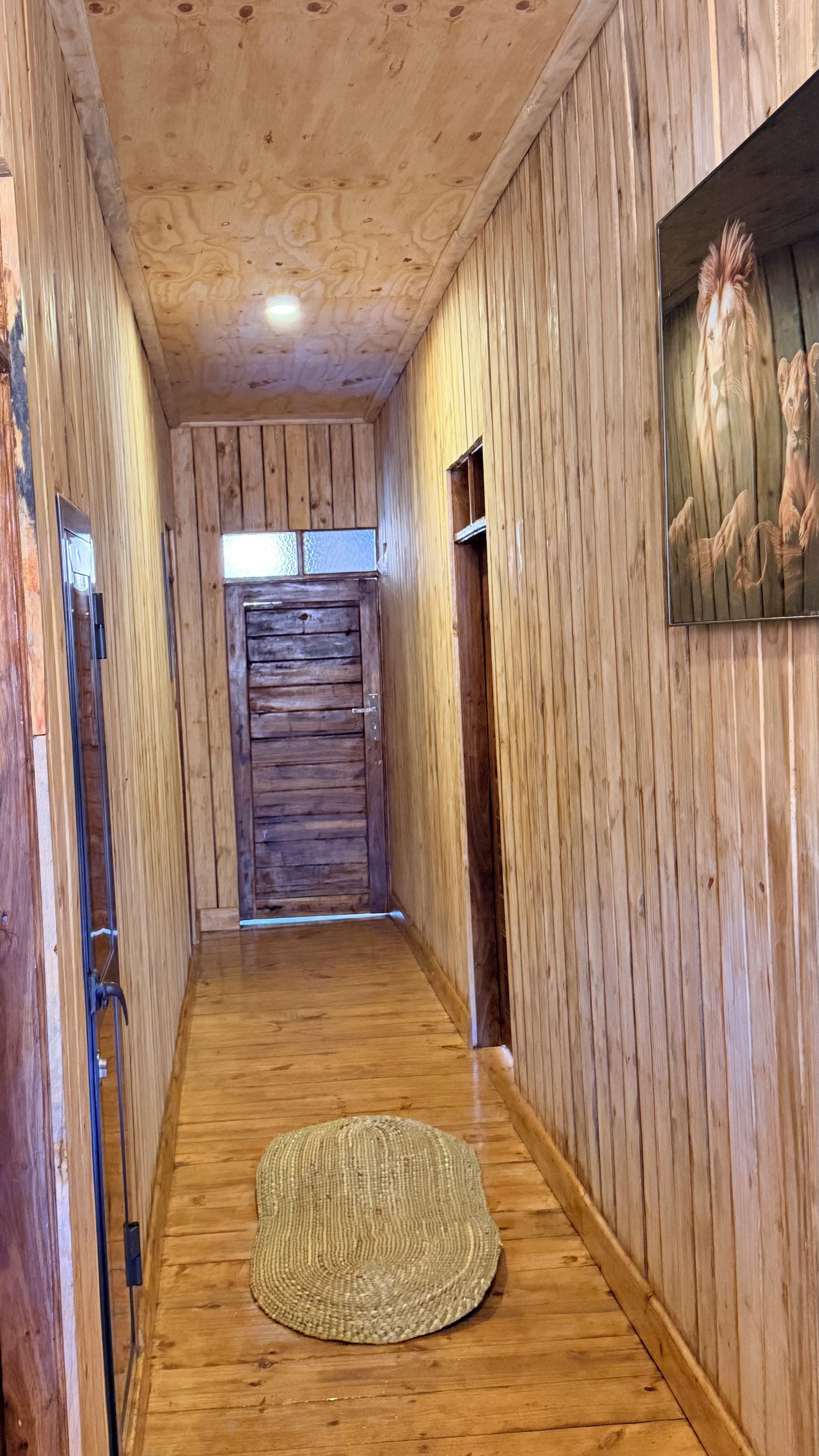 Um corredor de madeira com um tapete tecido no chão, uma porta de madeira no final e um quadro pendurado na parede.