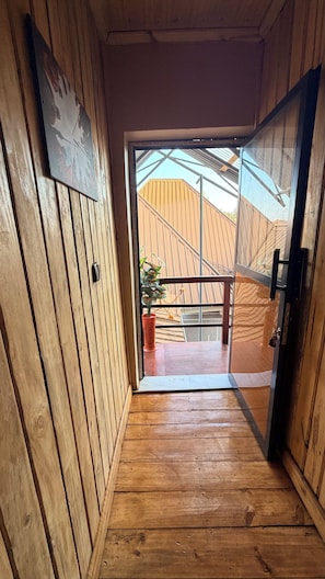 Passadís interior de fusta que condueix a una porta de vidre amb vista a una construcció de fusta a l'exterior.