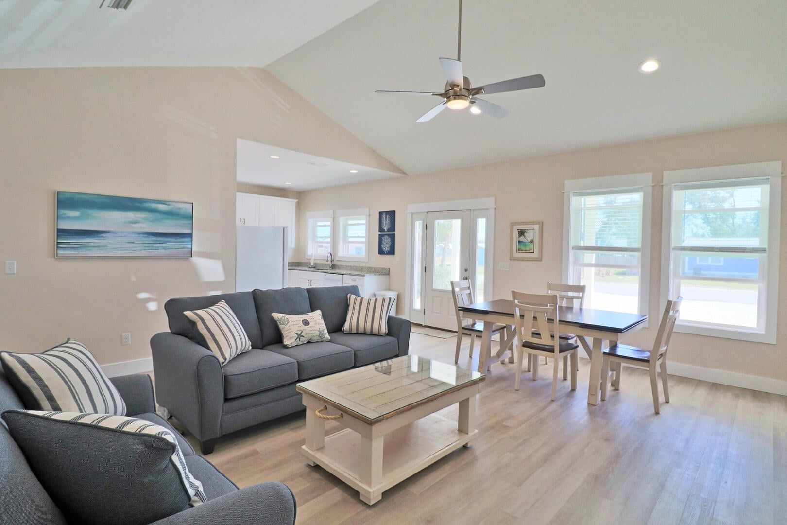A spacious living area with a gray sofa, a coffee table, and a dining table with chairs. There is a ceiling fan and a view of the outside through large windows.