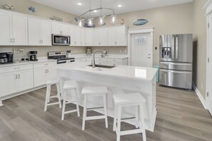 A modern kitchen with white cabinetry, a central island with bar stools, a stainless steel refrigerator, and a coffee bar. - THE "CRABBY HUNTER" IS A NEW HOME W/GULF VIEWS, PRIVATE POOL & OPTION TO RENT GO (Mexico Beach)