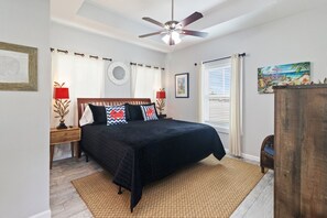 A bedroom with a bed, a ceiling fan, a nightstand with a lamp, a window with blinds, and a painting on the wall. - "Georgia Sand" is 600 steps to the beach! This 2BD/2BA adorable cottage sleeps 8 (Mexico Beach)