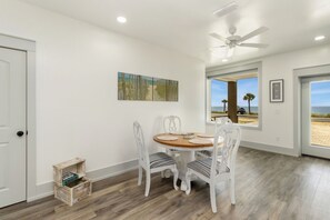 Ein Essbereich mit einem runden Tisch und weißen Stühlen, einem Deckenventilator und Blick auf den Strand durch große Fenster.