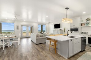 Cozinha moderna com ilha central, área de jantar com vista e sala de estar com sofá e poltronas.