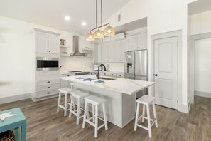 A modern kitchen with white cabinetry, a central island with bar stools, stainless steel appliances, and a hanging light fixture. - New Listing, Grand Coral III has gulf views & 100 steps to the beach! 2BD/1BA lu (Mexico Beach)
