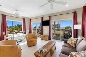 Ein großzügiger Wohnbereich mit Deckenventilator, einer Couch, einem Sessel, einem Couchtisch und Blick auf den Strand durch große Glaswände.