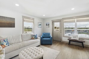 Een woonkamer met een bank, een blauwe fauteuil, een rond hocker en een bankje met een kussen.