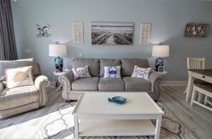 A living room with a grey sofa, two side tables with lamps, a coffee table, and decorative wall art. - PEACEFUL EASY FEEELING OFFERS GULF VIEWS & STEPS FROM BEACH! COMMUNITY POOL! (Mexico Beach)