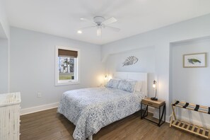 Ein Schlafzimmer mit einem Bett, einem Deckenventilator, einem Nachttisch mit Lampe, einem Fenster mit Aussicht und einem an der Wand befestigten Regal.