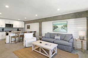A modern kitchen with white cabinets and stainless steel appliances, a dining area with wooden chairs, a grey sofa with a cushion, and a painting of a coastal scene on the wall. - "Salt Therapy" offers gulf views, 100 steps to beach, modern 2BD/2BA, single lev (Port Saint Joe)