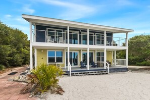 Une maison de deux étages située en bord de mer, dotée d’un balcon, d’un espace salon extérieur et d’une allée menant à la maison recouverte de sable.