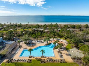Ett semesterhotell med en stor pool omgiven av palmer och solstolar, med utsikt över en strand.