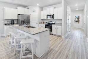A modern kitchen with white cabinetry, a granite countertop island with bar stools, stainless steel appliances, and a view into the adjacent living area. - SEA SMILE offers amazing sunsets, amenities galore & mother-in-law suite! Snowbi (Port Saint Joe)