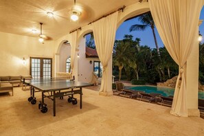 A ping pong table in a room with a pool, a sofa, and curtains. - Luxury Mansion | 7K SF | Pool & Game Room (Plantation)