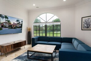 A living room with a sectional sofa, a coffee table, a TV mounted on the wall, and a large window with a view of a house and greenery. - Luxury Mansion | 7K SF | Pool & Game Room (Plantation)