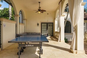 Taula de ping-pong a l'aire lliure amb un ventilador de sostre, situada davant d'un edifici amb arcs i una porta.
