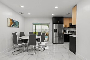 Une cuisine moderne équipée d’un réfrigérateur en acier inoxydable, d’une table à manger avec des chaises, et offrant une vue sur une piscine à travers des portes vitrées.