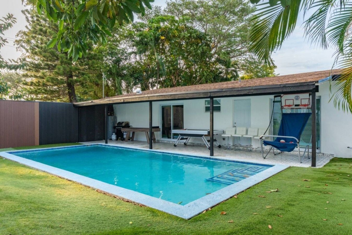 A swimming pool with a sunshade, a table, and chairs, surrounded by a fenced yard and trees.