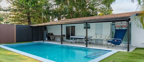 A swimming pool with a sunshade, a table, and chairs, surrounded by a fenced yard and trees.