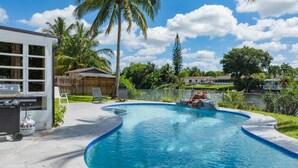 Una piscina con borde curvo, rodeada por una valla, palmeras y una casa al fondo.