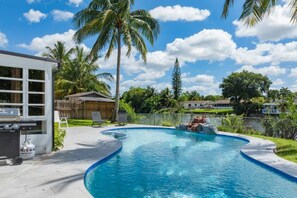 A swimming pool with a curved edge, surrounded by a fence, palm trees, and a house in the background. - 3‑BR Retreat w Pool near the Beach - Car charger (Pompano Beach)