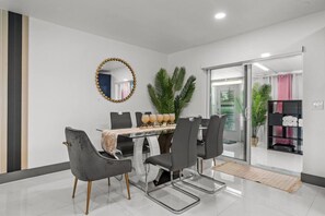A modern dining area with a glass table, grey upholstered chairs, and a decorative mirror. - 3‑BR Retreat w Pool near the Beach - Car charger (Pompano Beach)