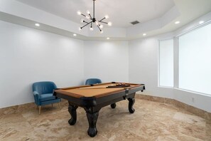 A pool table in a room with a modern ceiling light fixture, a blue chair, and a large window. - Luxury Mansion | 5K SF | Pool & Game Room (Hollywood)