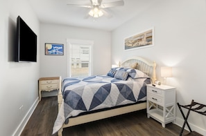 A bedroom with a bed, a nightstand, a bedside table, a ceiling fan, a TV mounted on the wall, and a window with blinds. - NEW LISTING! Special Sunset is steps to beach, hot tub, fenced backyard and pet (Mexico Beach)