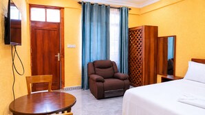 Habitación de hotel con una cama, un sillón marrón, una mesa de madera, un televisor montado en la pared y una ventana con cortinas.
