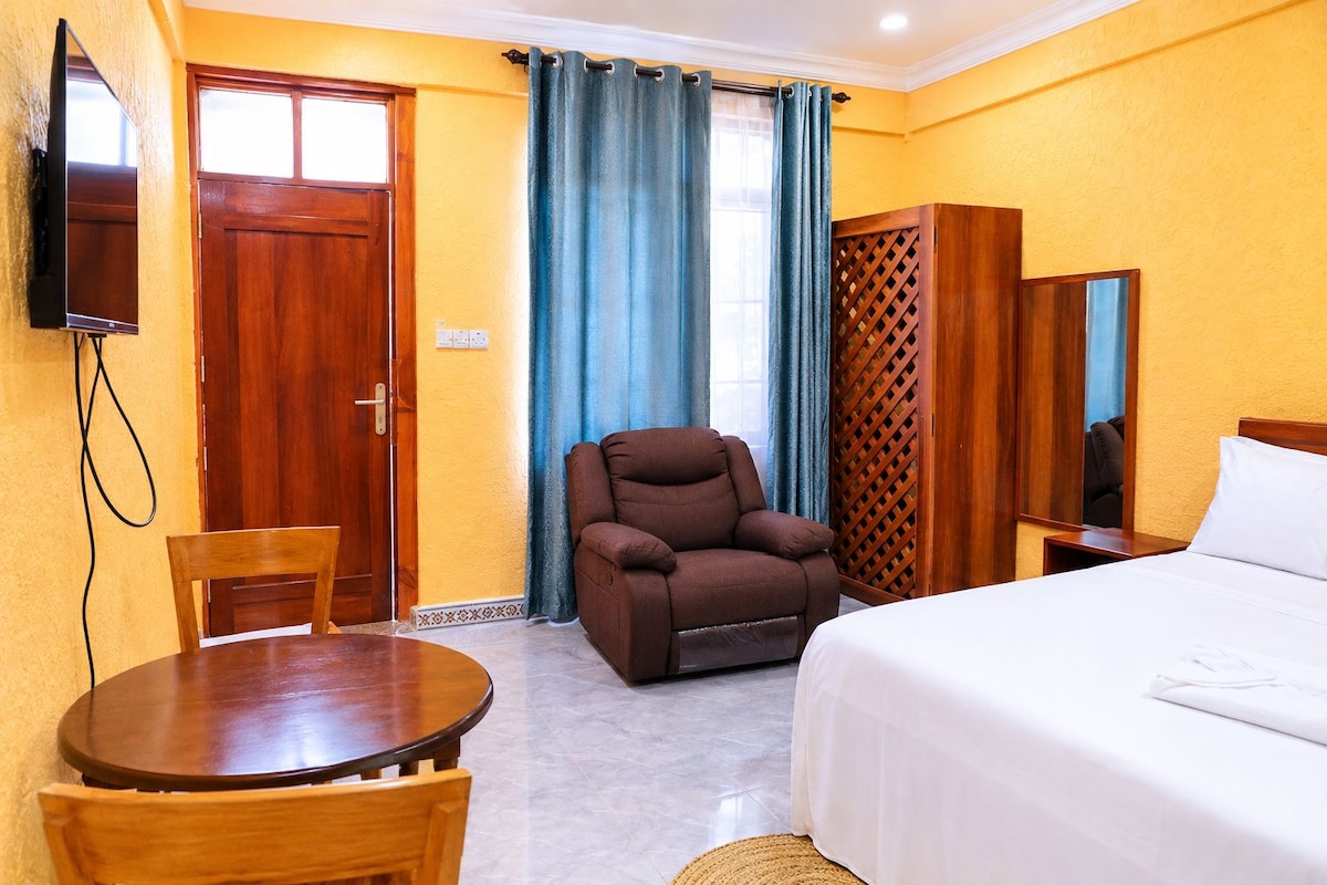 A hotel room with a bed, a brown armchair, a wooden table, a TV mounted on the wall, and a window with curtains.