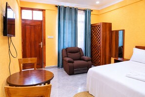 A hotel room with a bed, a brown armchair, a wooden table, a TV mounted on the wall, and a window with curtains. - BEST POINT HOTEL BWEJUU (dongwe)