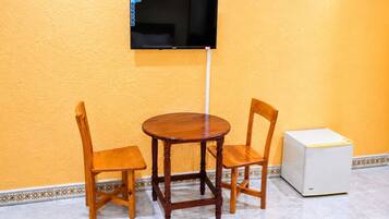 Una habitación con una mesa de madera y dos sillas, un televisor montado en la pared y un electrodoméstico blanco en el suelo.