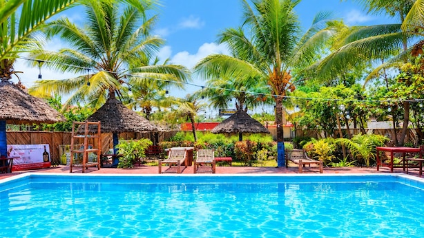 Una piscina azul cristalina rodeada de palmeras y cabañas con techo de paja, con sillones de descanso y una escalera de madera.