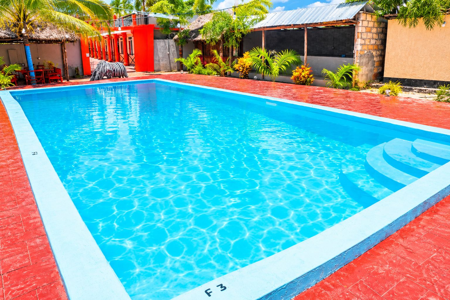 A rectangular swimming pool with clear blue water, surrounded by red and beige walls, and a small seating area with a zebra-patterned cushion.