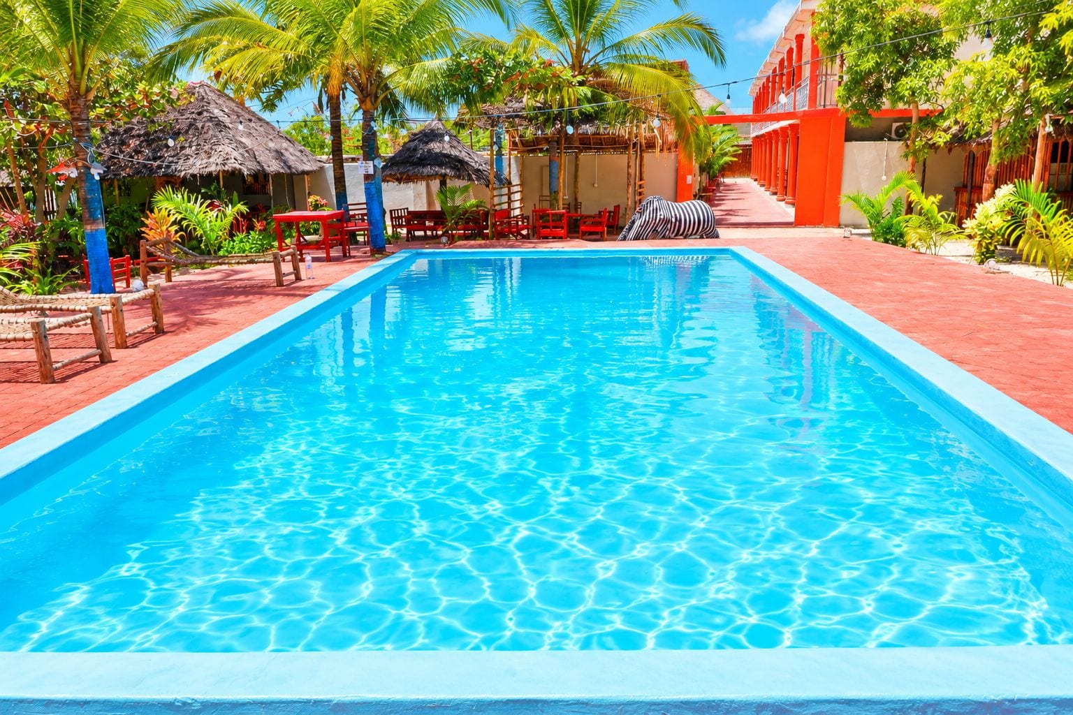 Una piscina con superficie de agua azul cristalina, rodeada de pavimento de ladrillo rojo, palmeras y estructuras con techo de paja.