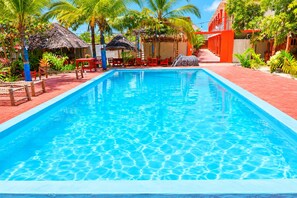 A swimming pool with a clear blue water surface, surrounded by red-brick flooring, palm trees, and thatched-roof structures.