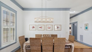 Un coin repas avec une table dressée pour quatre personnes, entourée de chaises en osier, d’un luminaire suspendu et de tableaux encadrés accrochés au mur.