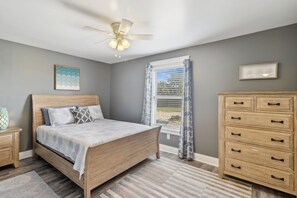 Ein Schlafzimmer mit einem Bett, einem Deckenventilator, einem Fenster mit Vorhängen, einem Schminktisch und einem Nachttisch.