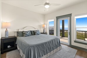 Ein Schlafzimmer mit einem Bett, Nachttischen, Lampen, einem Deckenventilator und Blick auf den Ozean durch eine Tür.