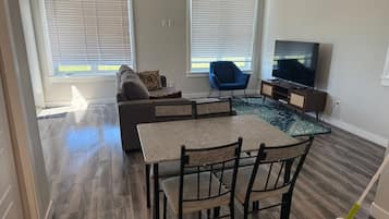 A living room with a sofa, a blue chair, a coffee table, a TV, and a dining area with chairs and a table.