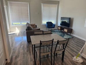 A living room with a sofa, a blue chair, a coffee table, a TV, and a dining area with chairs and a table.