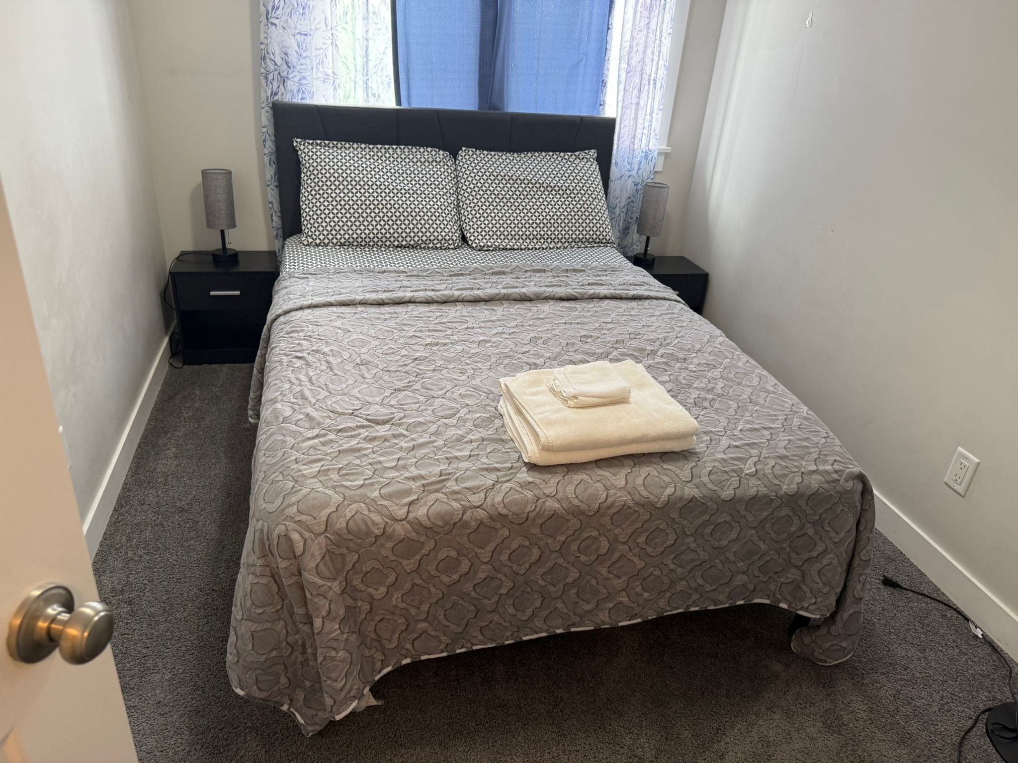 A neatly made bed with a patterned bedspread and pillows, a folded towel on the bed, and a nightstand with a lamp beside it.