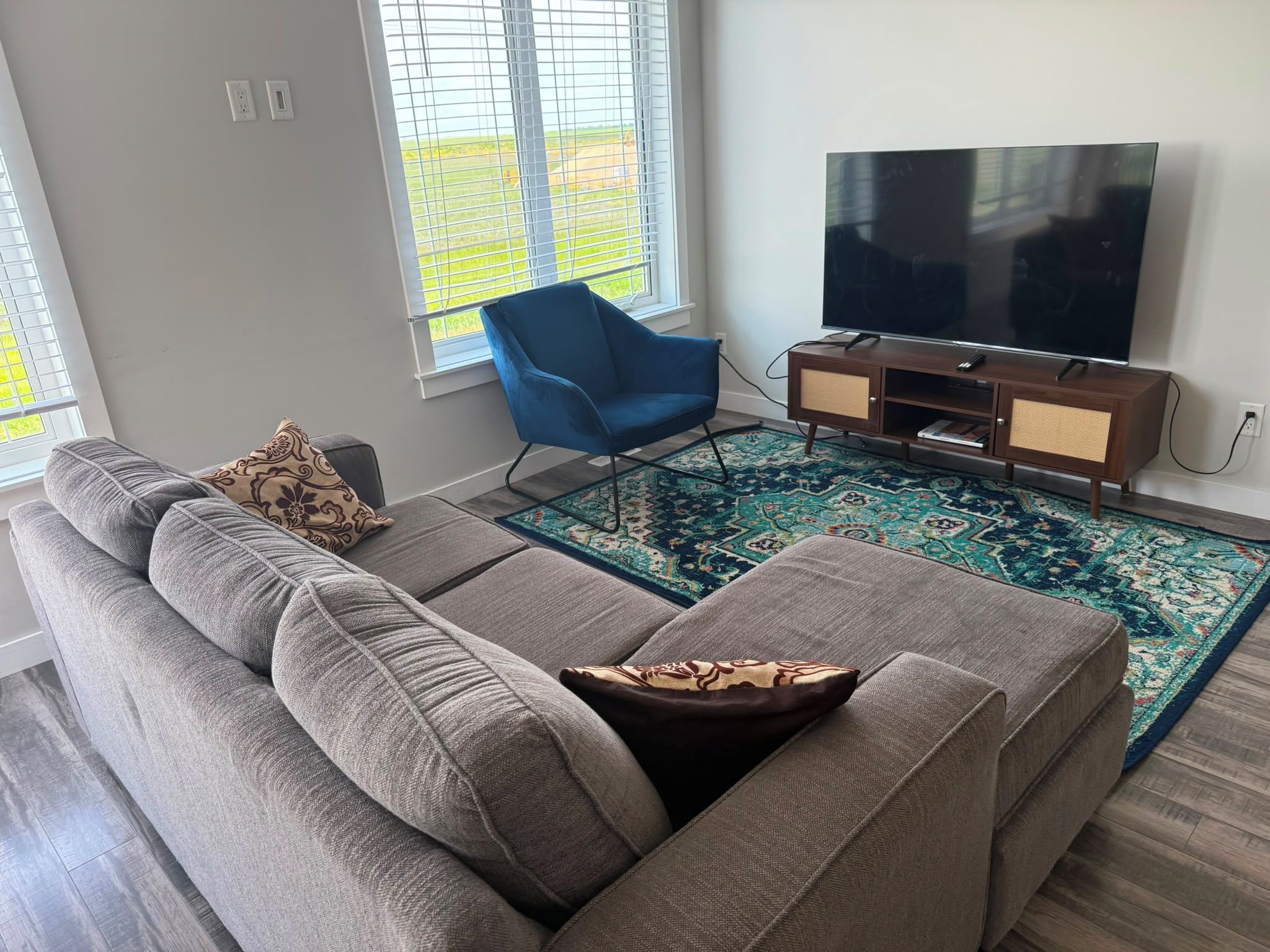 Un salon moderne avec un canapé d’angle gris, un fauteuil bleu, un téléviseur à écran plat posé sur un meuble en bois et un tapis à motifs.