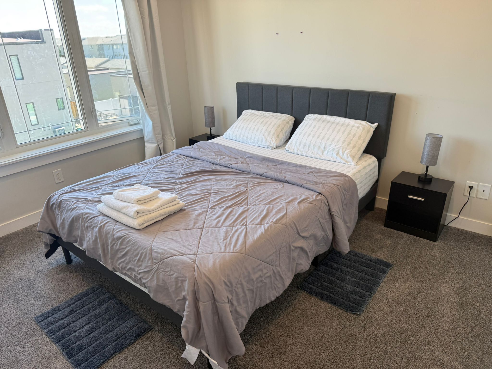A neatly made bed with a grey quilt and pillows, a small bedside table with a lamp, and a window with curtains.
