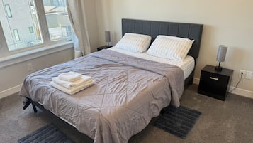 A neatly made bed with a grey quilt and pillows, a small bedside table with a lamp, and a window with curtains.