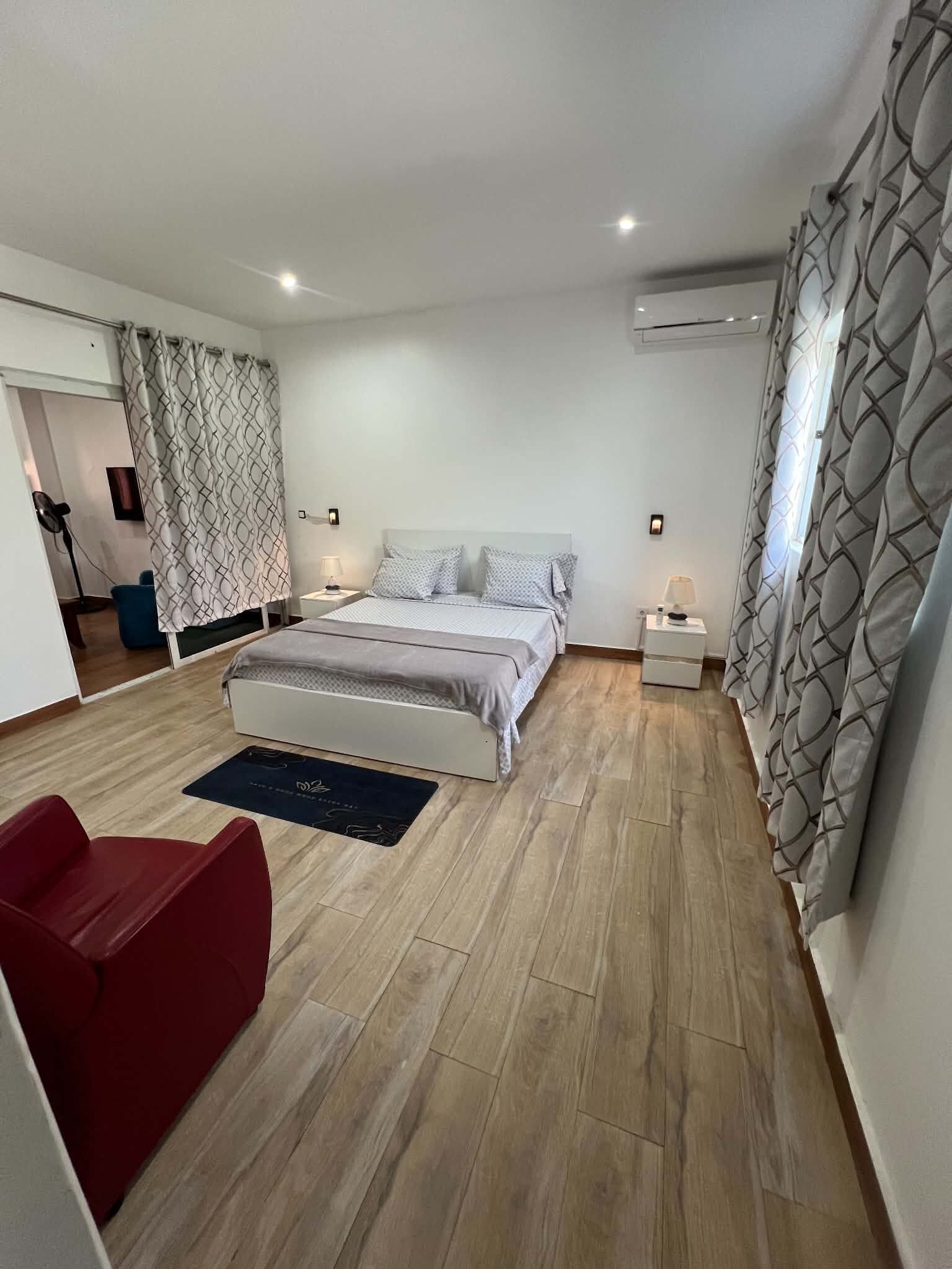 A bedroom with a bed, bedside tables, a red chair, a floor lamp, and patterned curtains.