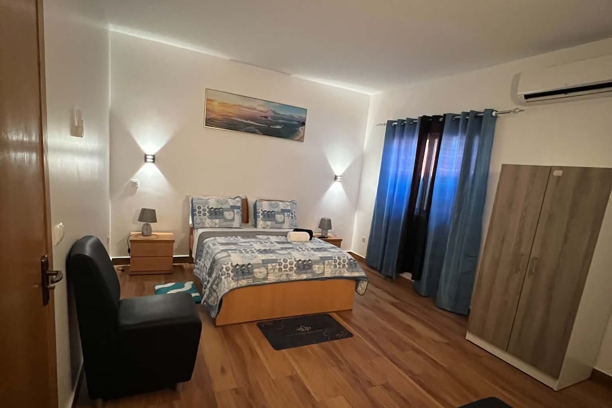 Un dormitorio con cama, mesitas de noche, una silla, armario y un cuadro en la pared.