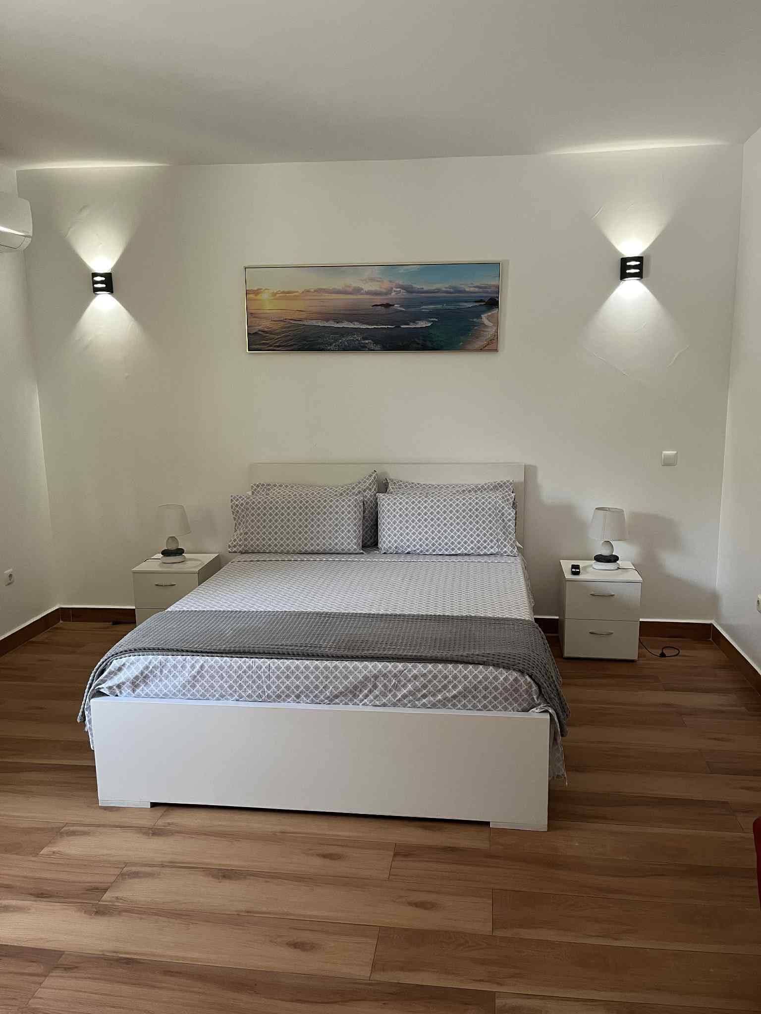 A bedroom with a bed, two bedside tables, a nightstand, a wall-mounted painting, and two wall-mounted lights.