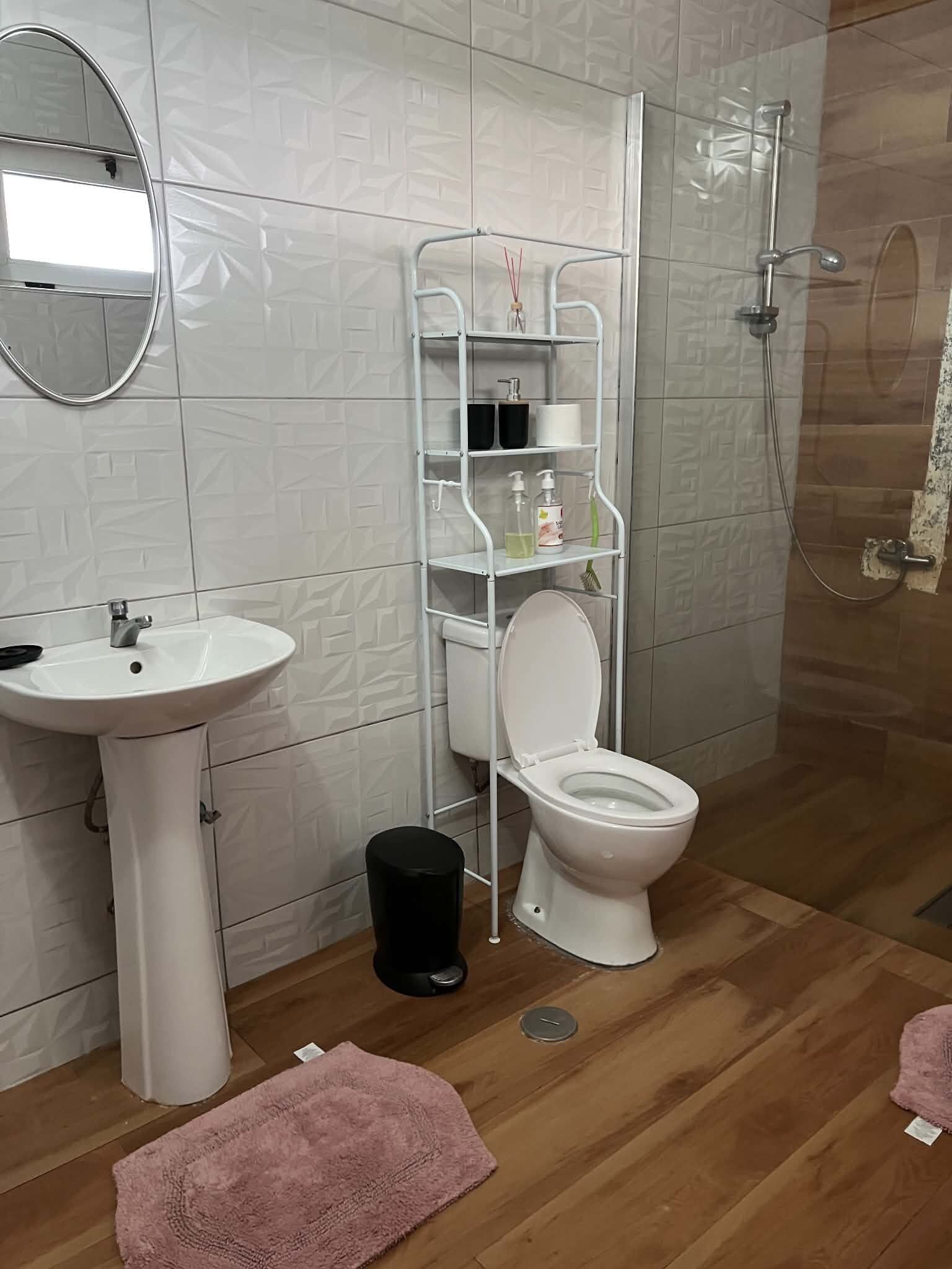 A bathroom with a white sink, toilet, and shower. There is a white shelf unit with toiletries.