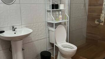 A bathroom with a white sink, toilet, and shower. There is a white shelf unit with toiletries.