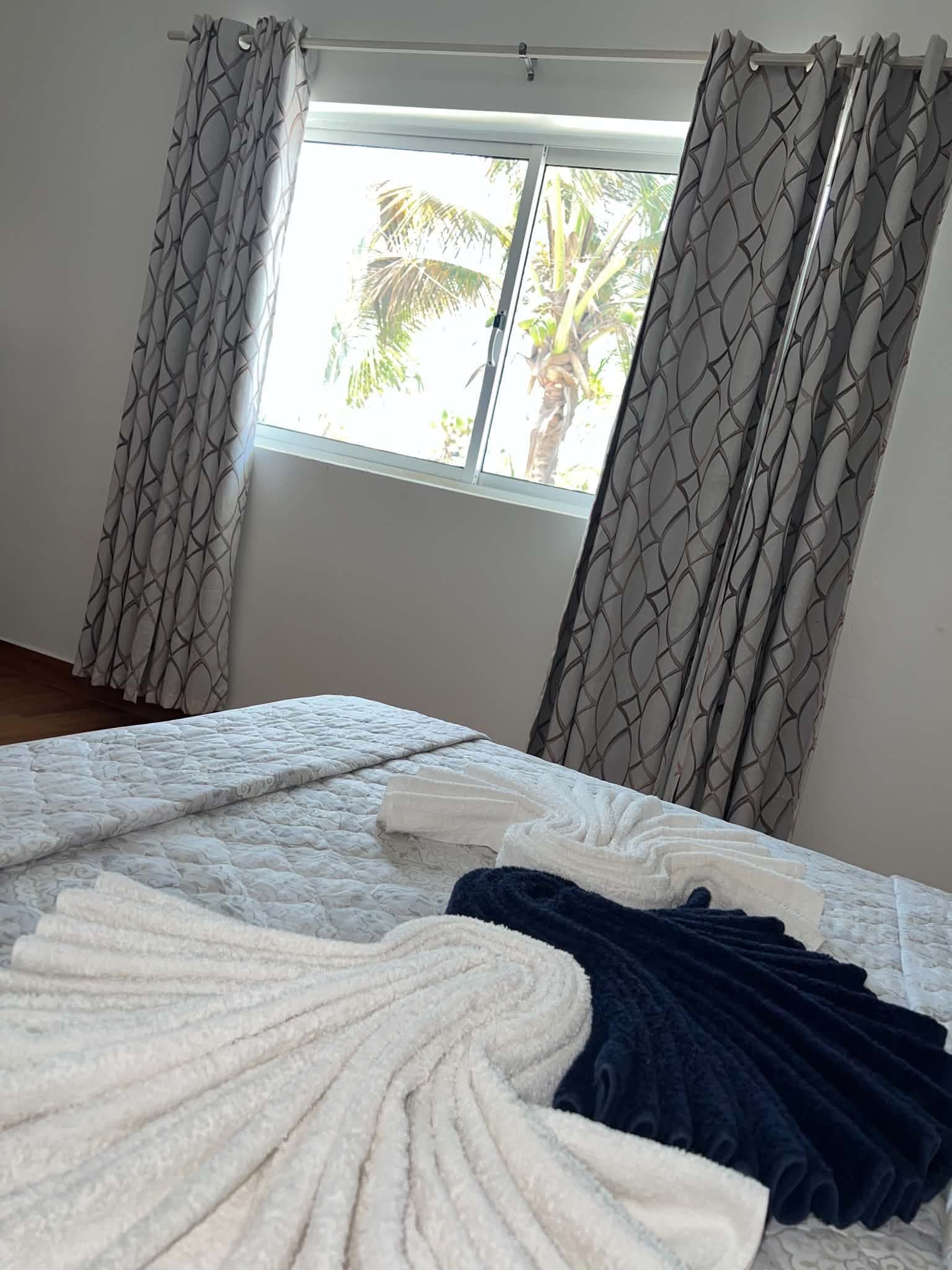 A bedroom with a bed, curtains, and a window with a view of palm trees.